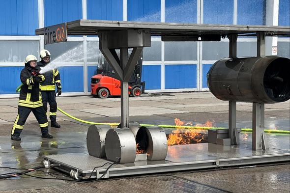 Zwei Feuerwehrleute spritzen Wasser auf die Brandsimulationsanlage